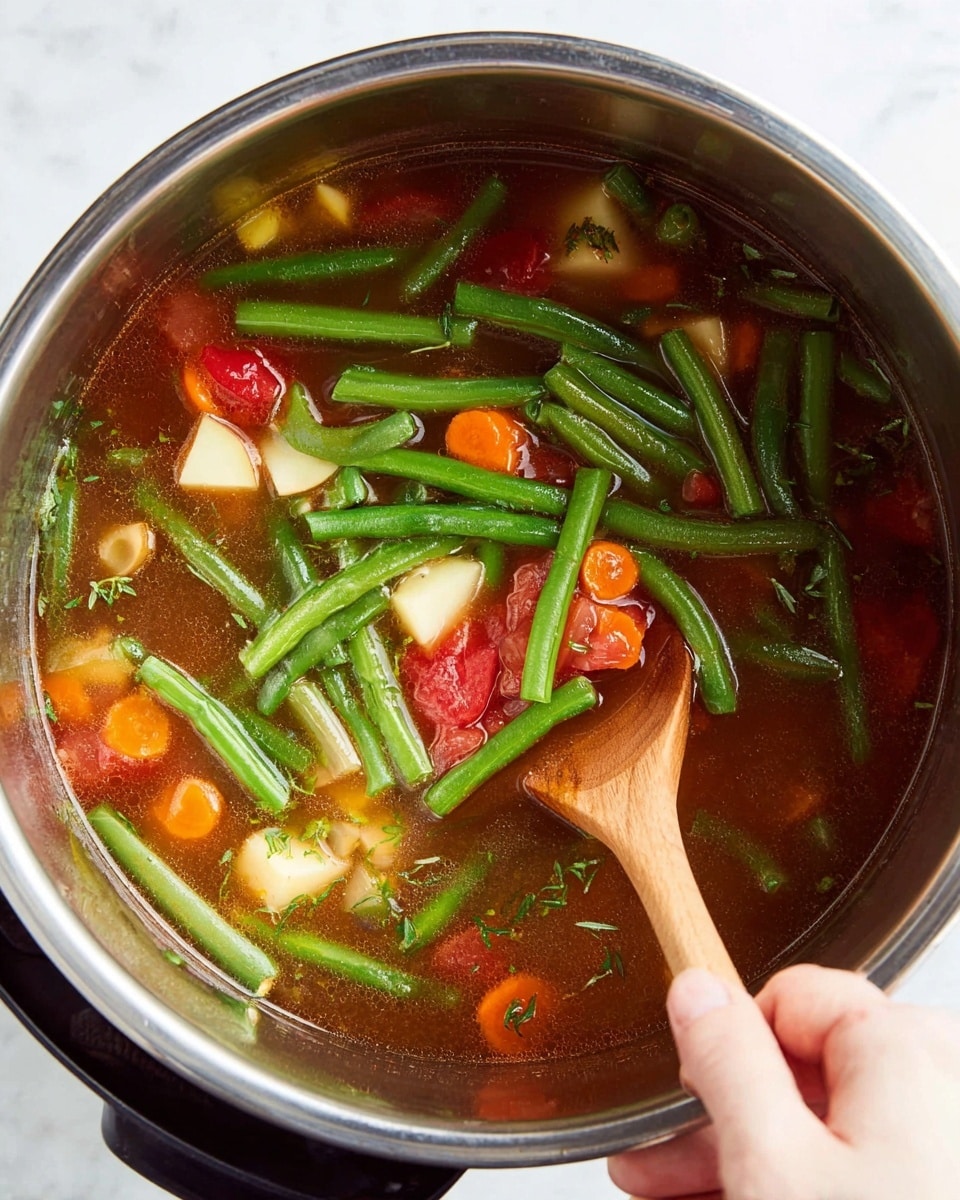 A close view inside a silver metal pot filled with clear brown broth soup, chunky with bright green whole green beans spread on top, with medium orange carrot slices, small white potato cubes, and large red tomato pieces mixed in. A woman's hand holds a wooden spoon inside the pot, stirring the ingredients, showing the details of the soup's herbs and vegetables. The pot sits on a white marbled surface, and light reflects softly on the metal rim photo taken with an iphone --ar 4:5 --v 7