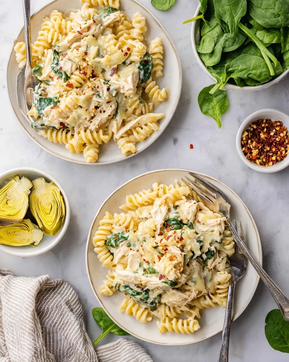 Two white plates filled with creamy pasta made of spiraled noodles mixed with shredded light-colored chicken and green spinach leaves, topped with melted light yellow cheese and tiny red pepper flakes. Each plate has a silver fork partially placed in the pasta. Next to them, there are small white bowls, one filled with bright yellow artichoke hearts and another with a small amount of red pepper flakes. Fresh green spinach leaves are scattered on a white marbled surface around the dishes, and a neutral striped cloth is partly visible at the side. photo taken with an iphone --ar 4:5 --v 7