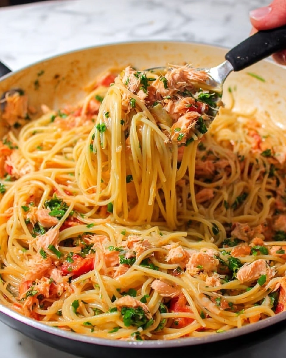 The image shows a close-up view of a large white pan filled with cooked spaghetti mixed with small chunks of pinkish-orange fish, chopped green herbs, and small red tomato pieces, all coated in a light sauce. The spaghetti strands are light yellow and appear soft and slightly shiny. A woman's hand holds a metal spoon with a black handle lifting a bunch of spaghetti from the pan, with strands dangling and some herbs and tomato bits clinging to the pasta. The background has a white marbled surface. Photo taken with an iphone --ar 4:5 --v 7