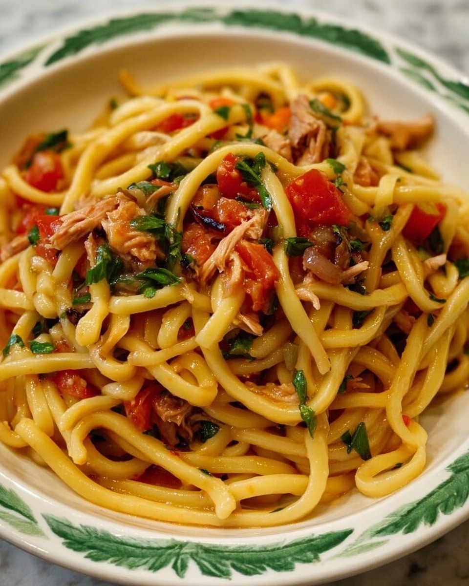 A close-up of a white bowl filled with a single layer of long, thick yellowish noodles twisted and mixed with chunks of red tomatoes, small pieces of light brown shredded meat, purple onion bits, and green herbs scattered throughout. The bowl has a green leaf pattern on the edge and sits on a white marbled surface. The noodles look soft with a slight shine from light sauce coating them. photo taken with an iphone --ar 4:5 --v 7