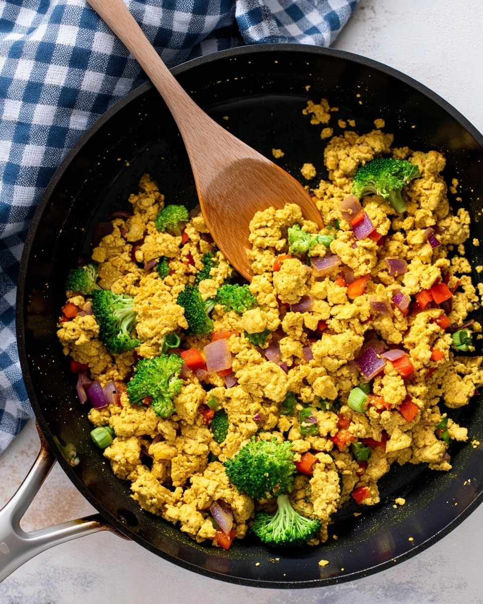 A black pan filled with a cooked mixture of crumbled yellow tofu as the main layer, mixed with small bright green broccoli florets, tiny red bell pepper pieces, and purple onion bits spread evenly throughout. A wooden spoon is partly resting on the mixture in the pan. The pan is placed on a surface with a white marbled texture and a blue and white checkered cloth peeks from the top left corner. Photo taken with an iphone --ar 4:5 --v 7