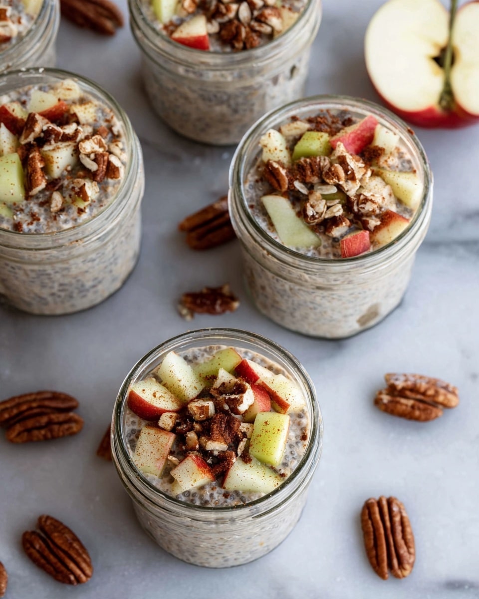 The image shows several clear glass jars filled with a creamy beige mixture that has small dark chia seeds and oats spread throughout. On top of each jar, there is a layer of small, fresh apple pieces with red and green skin, along with chopped pecans. A light sprinkle of cinnamon powder is visible over the fruit and nuts. The jars are placed on a white marbled surface, with scattered pecan halves and a halved apple near the jars. The photo taken with an iphone --ar 4:5 --v 7