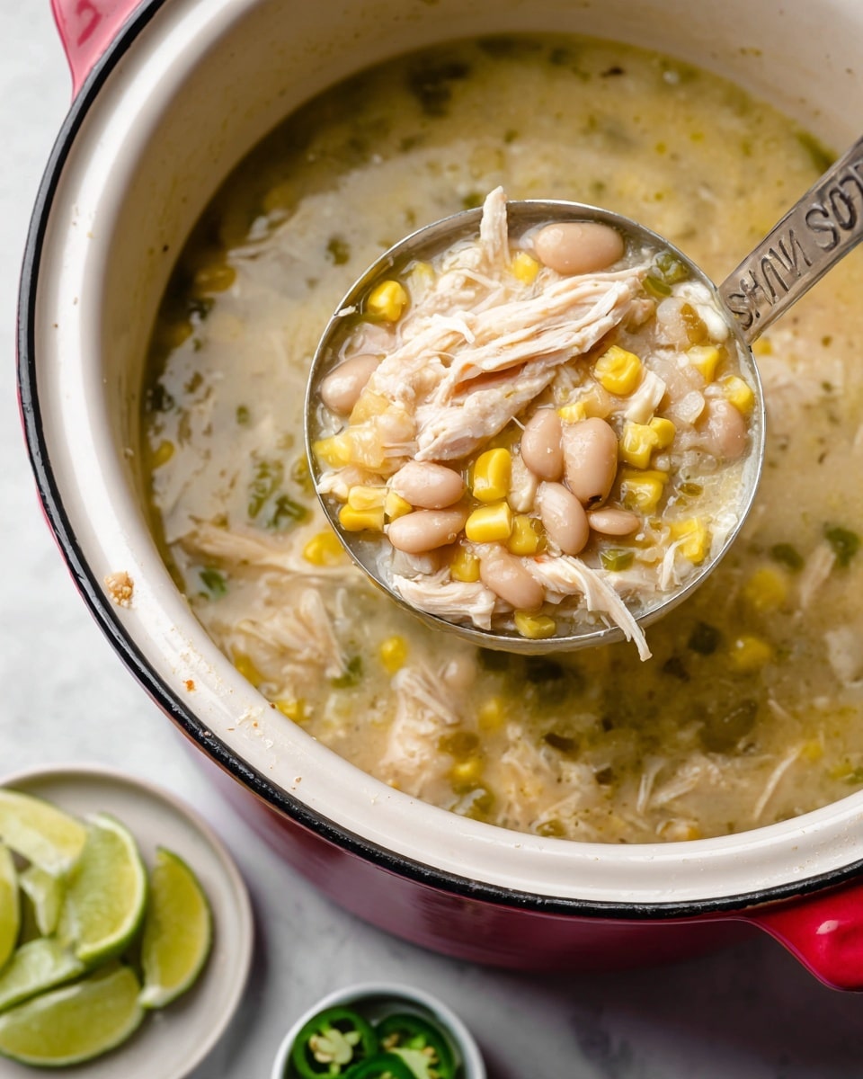A close-up of a metal ladle with shiny silver color holding thick soup made of several layers: shredded white chicken pieces, light yellow whole corn kernels, small white beans, small green chili pieces, and a light creamy soup base with a slightly chunky texture. The ladle is inside a white crock pot with a shiny red rim and edges. The background shows a clear jar with a white lid and a green and yellow label on a white marbled surface, along with lime slices on a plain white plate and small green chili pieces in a small white bowl. Photo taken with an iphone --ar 4:5 --v 7