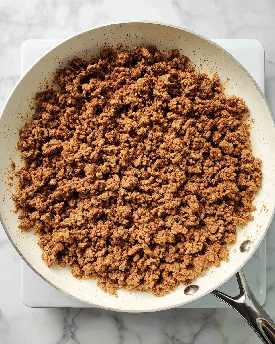 A white frying pan filled with evenly cooked ground beef, showing a layer of small, crumbly brown meat pieces spread out flat across the entire pan. The beef has a slightly crumbly texture with a mix of darker and lighter brown shades throughout. The pan sits on a stove with silver handles visible, and the background is a white marbled surface. photo taken with an iphone --ar 4:5 --v 7