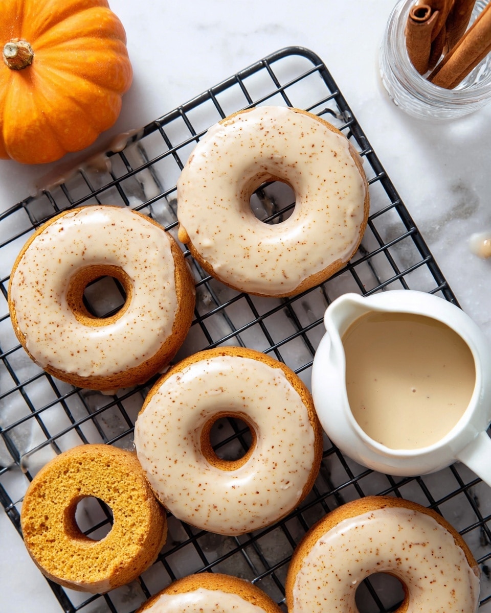 The image shows seven pumpkin-colored donuts with a soft texture, six of them topped with a smooth, light beige glaze sprinkled with small brown specks, and one donut without glaze, revealing its spongy inside. The donuts are placed on a black cooling rack resting on a white marbled surface. To the right, there is a white small pitcher filled with the same glaze and a silver spoon inside. At the top left corner, there is a small bright orange pumpkin, and at the top right corner, a clear glass with two cinnamon sticks inside. The photo is bright and clear, taken from above, capturing all the details of the donuts and the surrounding items photo taken with an iphone --ar 4:5 --v 7