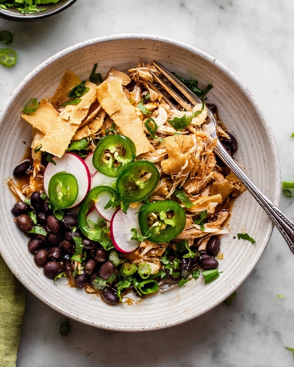 In a white bowl with a subtle textured edge, there is a layered dish starting with shredded light brown cooked chicken at the base, topped with black beans scattered throughout. On top, there are golden crispy tortilla strips arranged in a scattered manner, with thin slices of bright green jalapeño peppers and white radish slices with red edges placed beside and on the tortilla strips. Fresh chopped green herbs and green onions are sprinkled across the dish. A silver fork is placed inside the bowl on the left side, resting against the food. The bowl is set on a white marbled surface with some sliced green jalapeños scattered nearby. photo taken with an iphone --ar 4:5 --v 7
