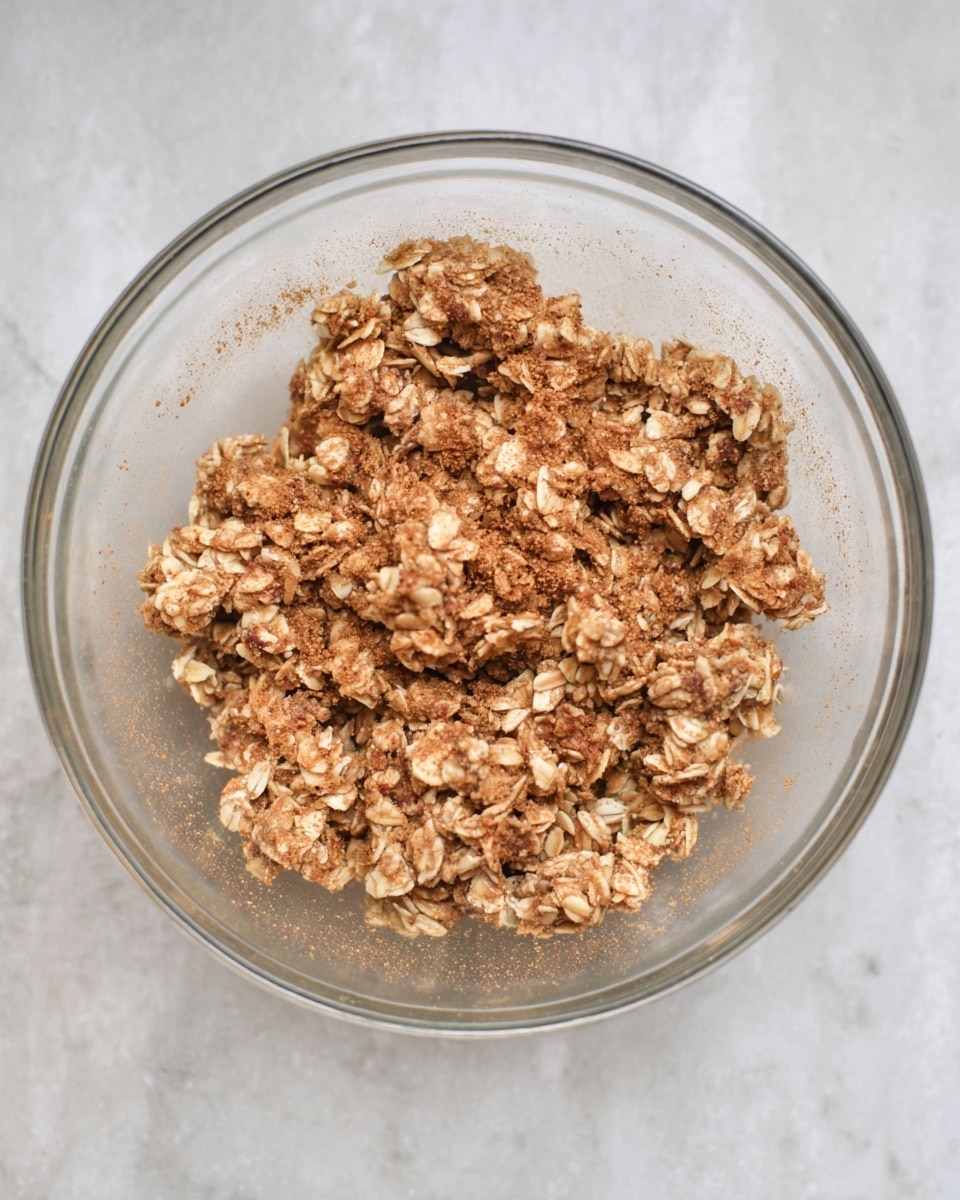 A clear glass bowl holds a mix of crumbled granola with visible oats and bits of nuts or seeds. The granola is light and medium brown, giving a rough texture with some small clusters. The inside of the bowl has a light dusting of cinnamon or spice powder, adding a soft orange-brown color coating. The bowl sits on a white marbled surface, giving a clean and bright background. Photo taken with an iphone --ar 4:5 --v 7