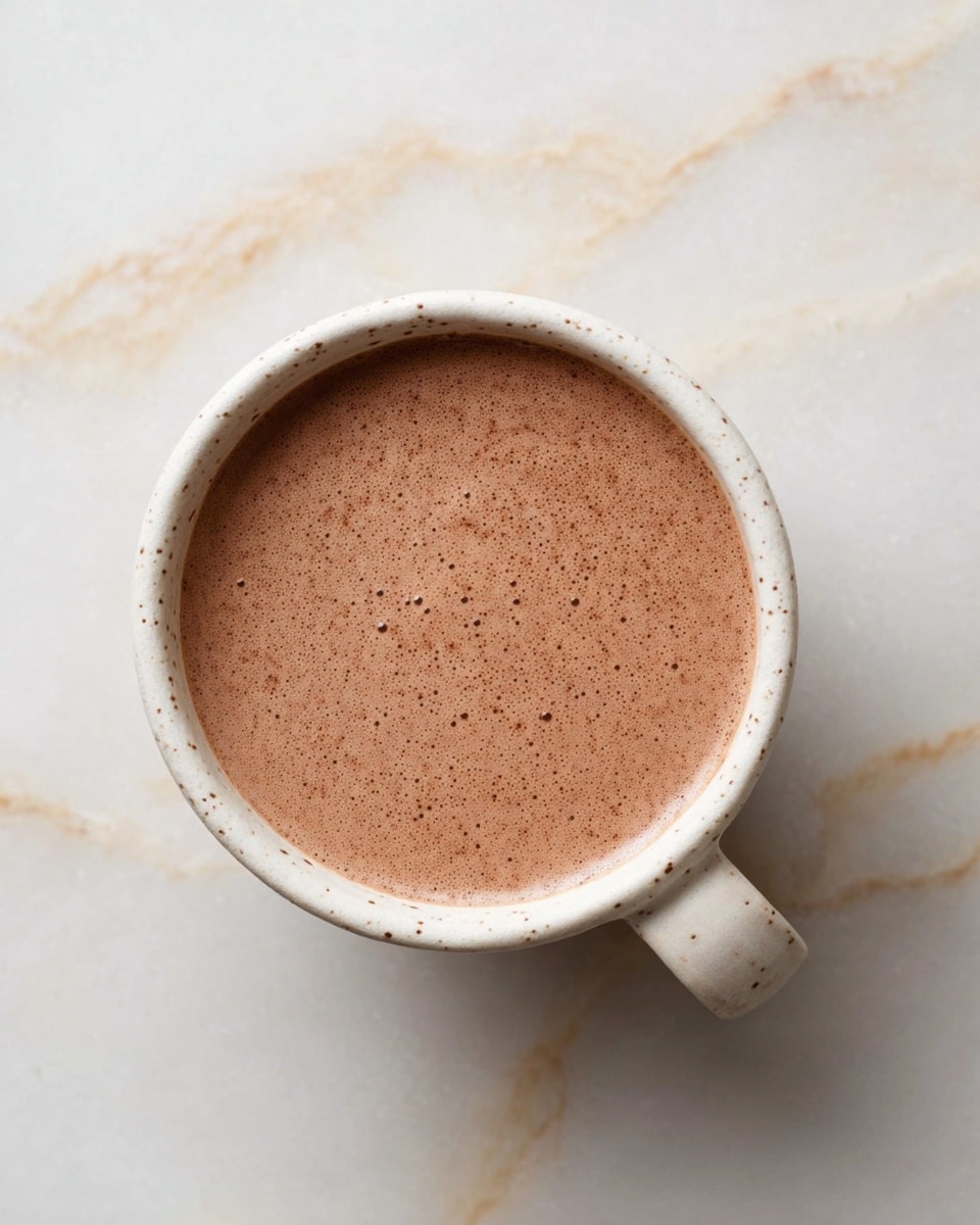 A top view of a white speckled ceramic cup filled with smooth, light brown hot chocolate without any toppings, placed on a white marbled surface with soft beige veins. The hot chocolate has a frothy texture with tiny bubbles spread evenly across the surface. The cup handle is visible on the right side, matching the cup’s speckled cream color. photo taken with an iphone --ar 4:5 --v 7