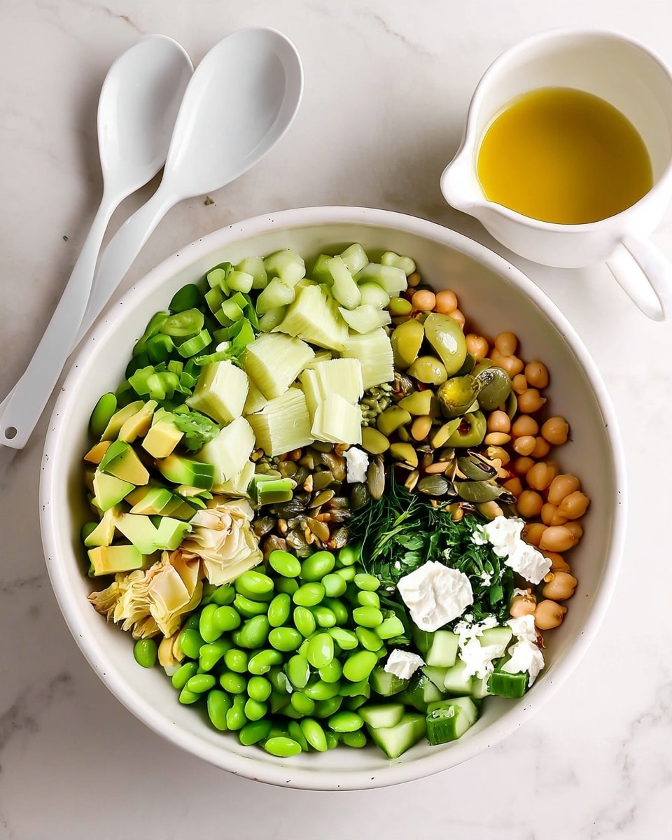 The image shows a white bowl filled with a colorful, fresh salad arranged in sections. Starting from the bottom left and moving clockwise, there are bright green edamame beans with a smooth texture, light green chopped artichoke hearts, light green diced avocado pieces, darker green chopped celery, green olive slices, a small pile of mixed green and brown pumpkin seeds, light tan chickpeas, diced cucumber with a fresh and moist look, crumbled white cheese scattered over the vegetables, and chopped fresh green herbs near the center. To the right of the bowl, there are two white spoons placed on a white marbled surface, and a small white cup with a spout filled with golden olive oil is partially visible in the top right corner. The photo taken with an iphone --ar 4:5 --v 7