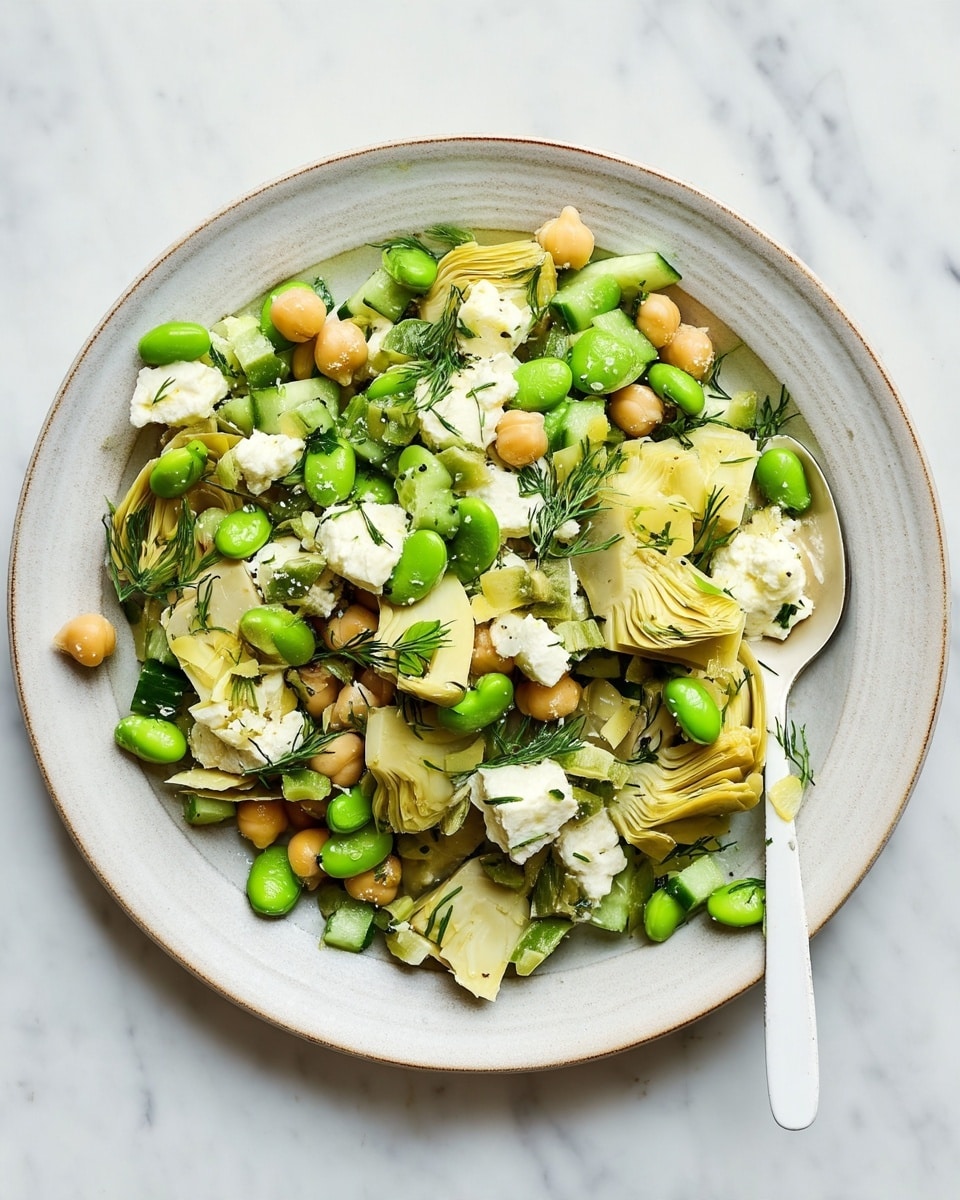 The dish is a salad served in a shallow, round white plate with a light, speckled texture. The salad has several visible layers starting with a base of pale yellow artichoke pieces that have a soft, slightly fibrous texture. On top of that are green edamame beans, adding a smooth and shiny texture. Scattered among the edamame and artichoke are creamy white chunks of feta cheese, which look crumbly and soft. There are also small, round beige chickpeas layered evenly throughout the salad. Small pieces of green cucumber and finely chopped fresh dill are mixed in, adding a fresh and slightly crunchy texture. The salad's overall look is colorful, fresh, and full of different textures. A white spoon is partially placed inside the plate on the right side against a white marbled surface. Photo taken with an iphone --ar 4:5 --v 7