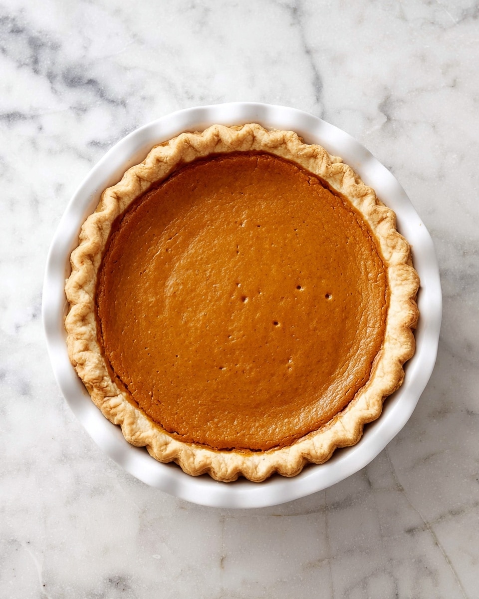 The image shows a single-layer pumpkin pie in a white pie dish. The pie has a golden-brown, smooth filling with a slightly glossy finish and a few small cracks. The crust is light beige with a crimped, scalloped edge that frames the filling neatly. The pie dish is placed on a round, white marbled textured surface. photo taken with an iphone --ar 4:5 --v 7
