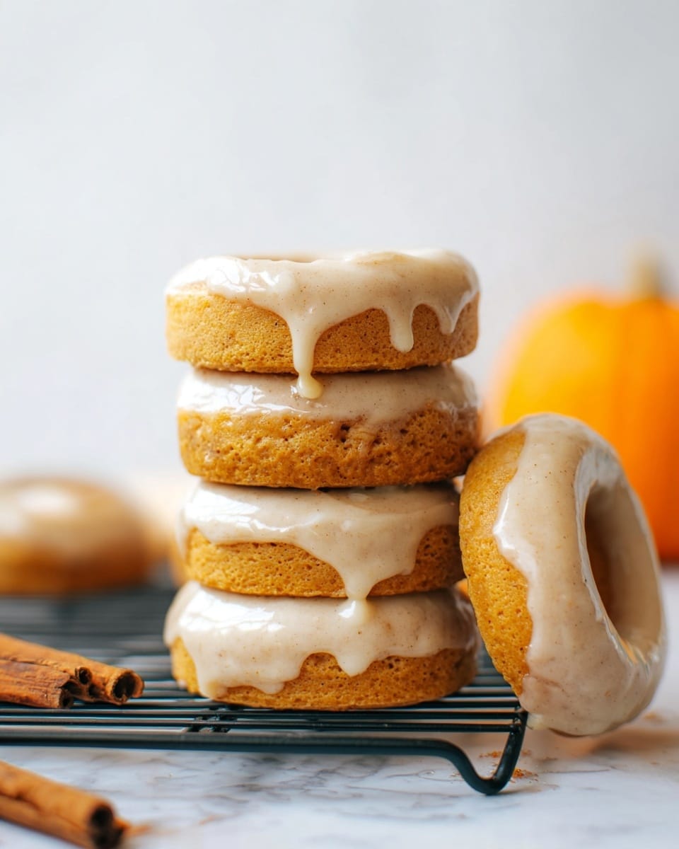 A stack of four round donuts with a light brown color sits on a black wire rack over a white marbled surface. Each donut is topped thickly with creamy, light beige icing that drips slightly down the sides. One more donut leans against the right side of the stack, similarly frosted. Near the bottom left corner, a cinnamon stick and a small section of another donut are visible, and in the blurred background, there is an orange pumpkin shape. The image has soft, natural light and a clean white background. photo taken with an iphone --ar 4:5 --v 7