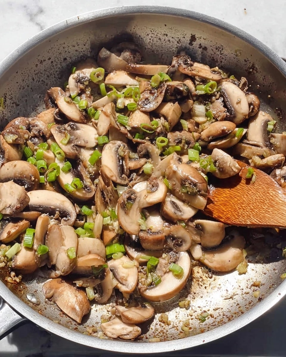 The image shows a silver frying pan filled with cooked mushroom slices and small bits of green onion. The mushrooms appear soft and slightly browned with a light beige inside and darker brown edges. The green onion pieces are scattered evenly on top, adding bright green color. There is a wooden spoon on the right side partly resting in the pan, mixing the mushrooms. The pan's surface around the food has small browned bits from cooking. The background is a white marbled texture. photo taken with an iphone --ar 4:5 --v 7